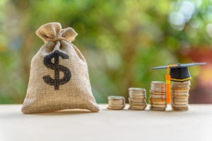 Education expense or student loan for post secondary education concept : Dollar bag, graduation cap on row of coins on a table, depicts loan or money designed to help students pay for associated fees