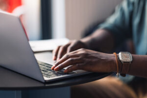 Close up photo of unrecognizable African-American man hands typing homework on his laptop keyboard while sitting in a coffee shop.