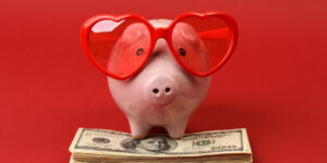 Piggy bank in love with red heart sunglasses standing on stack of money american hundred dollar bills on red background