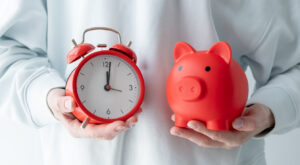 A person is holding a red piggy bank and a red alarm clock. The piggy bank is red and has a cute design on it